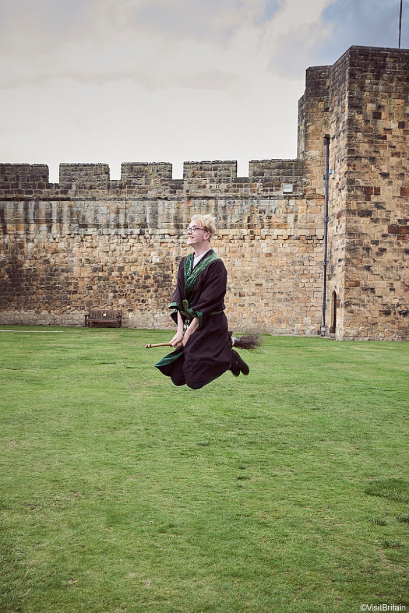 ©VisitBritain - Young man wearing robe and glasses hovering on broomstick in the Outer Bailey of Alnwick Castle, Northumberland